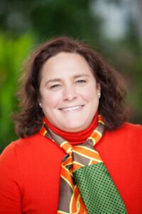 Photo of Kelly Velasquez-Hague wearing a red outfit with trees in the background. She is also wearing an orange and green scarf.