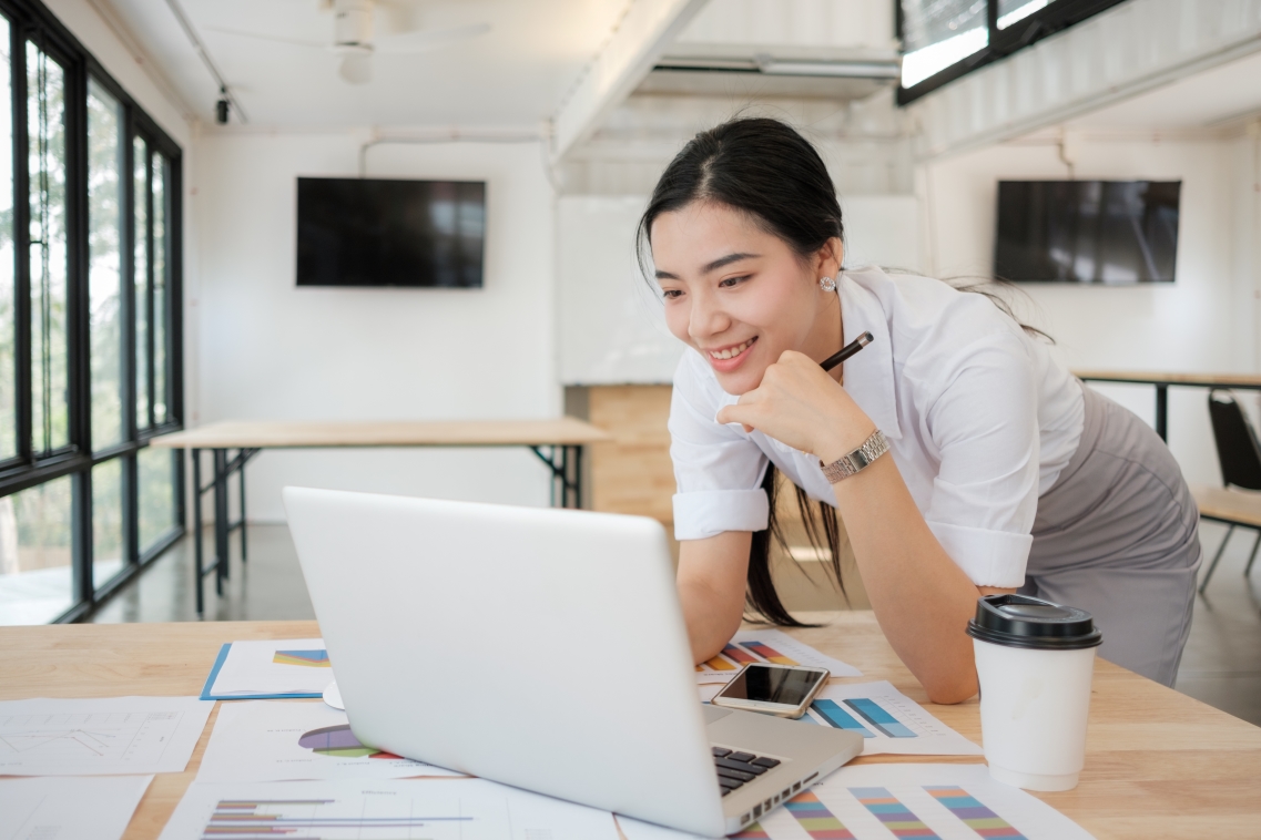 A woman in business casual attire leans over her desk and scrolls on her laptop